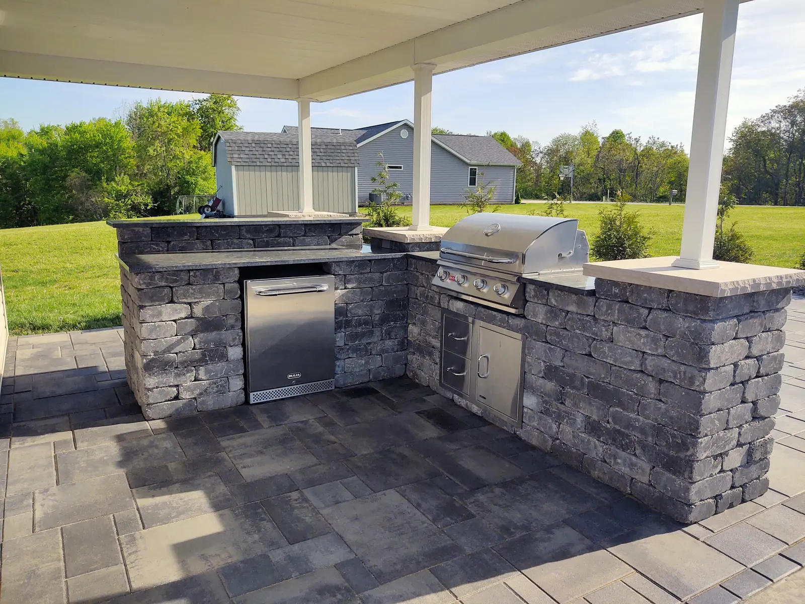 Stone outdoor kitchen and grill beneath a covered patio by Dave's Landscaping