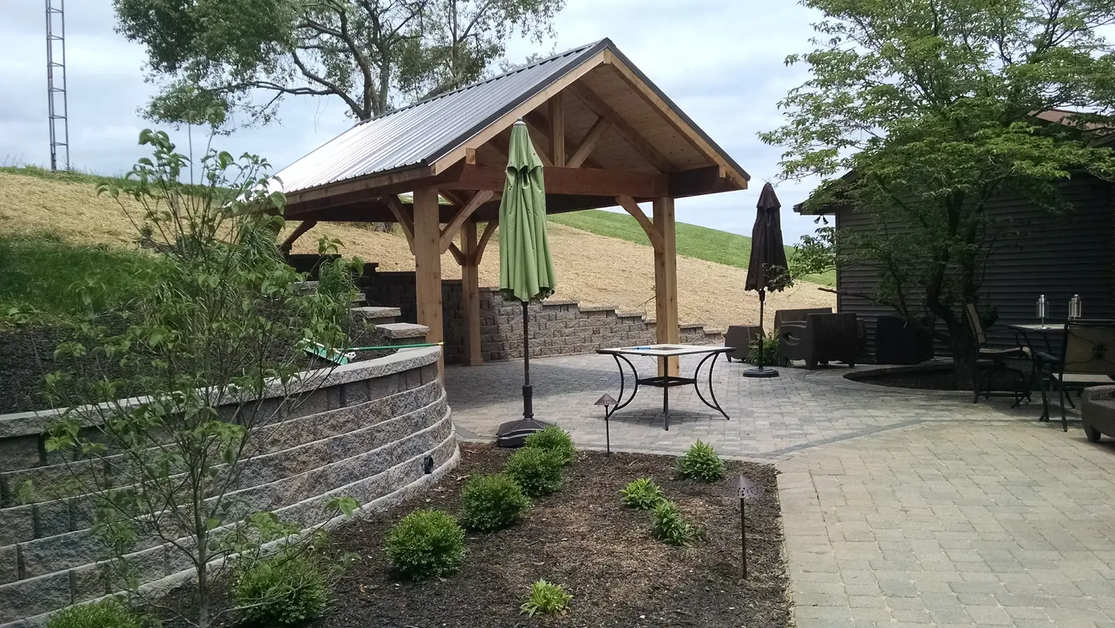 Paver patio and timber pavilion tucked into a landscaped slope by Dave's Landscaping